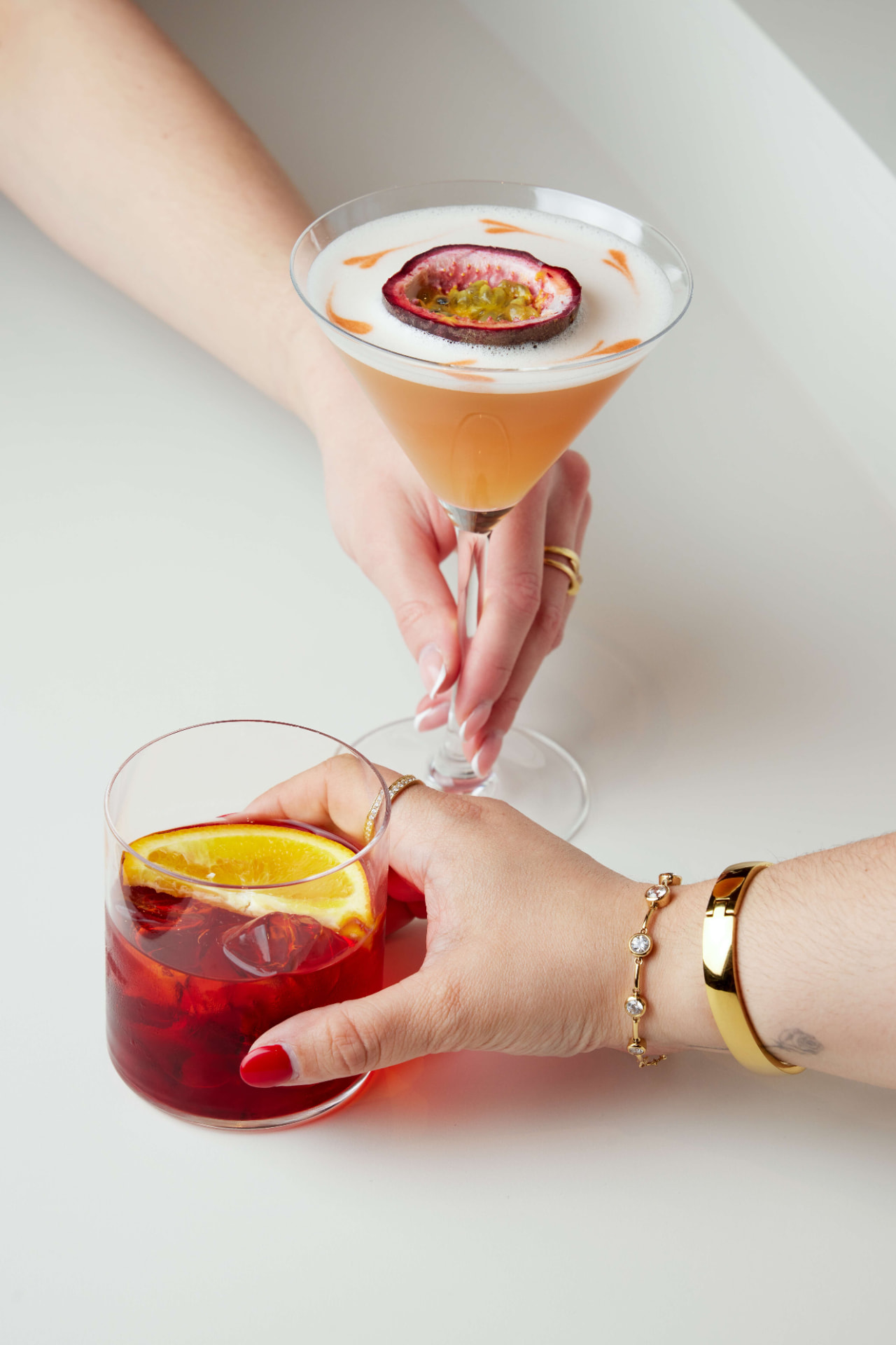 Zwei Hände halten einen Maracuja-Cocktail und einen Negroni über einem weißen Tisch